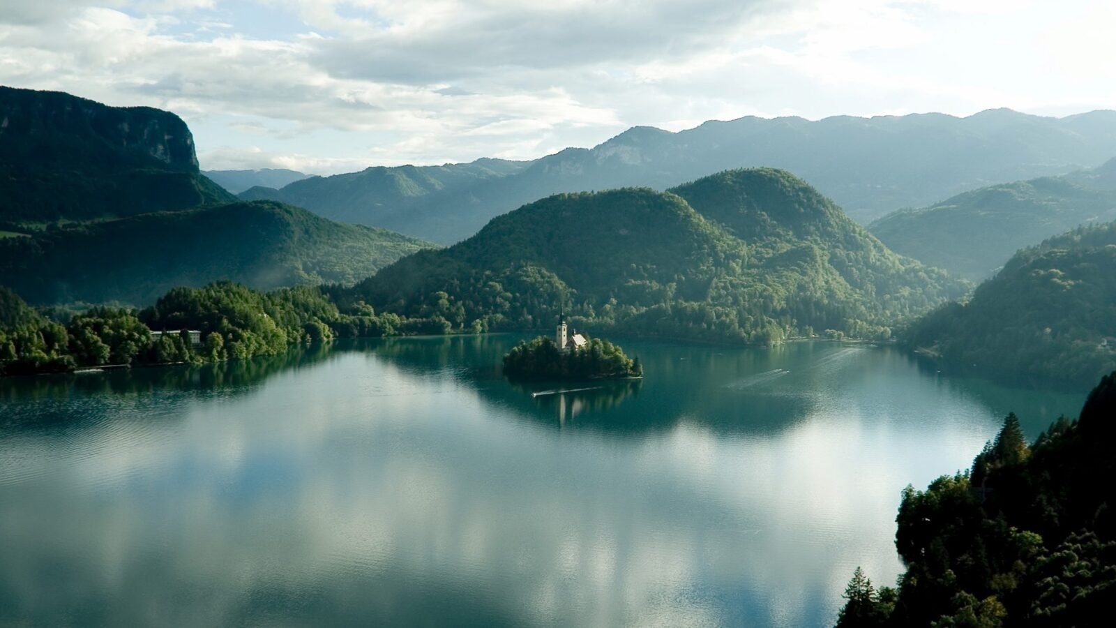 Aerial view of Lake Bled in the Alps, with a small church on an island in the middle of clear still lake surrounded by mountains and thick forests
