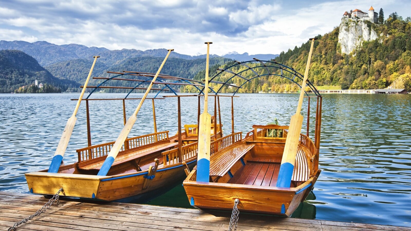 Two wooden open-top row boats mored by a wooden deck on Lake Bled in Slovenia. Beyond the boats you can see a castle high up on a cliffside and a small island in the water with a church on it. The lake is surrounded by forested mountains.