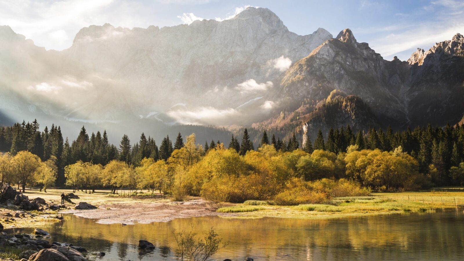 Early morning golden sunlight streaming over soft yellow and green trees by a still Lake Bled in Slovenia. The lake is surrounded by mountains which have a blanket of soft low clouds.