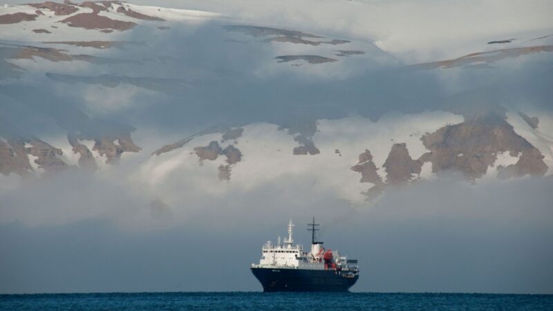 A white and black ship on the ocean with large, snowy, cloud-covered mountains in the distant background.