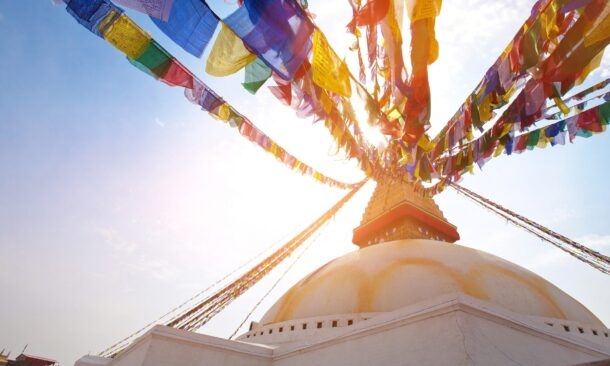 Sunlight shines through rows of colorful Buddhist prayer flags connected to the golden spire of a white domed stupa.