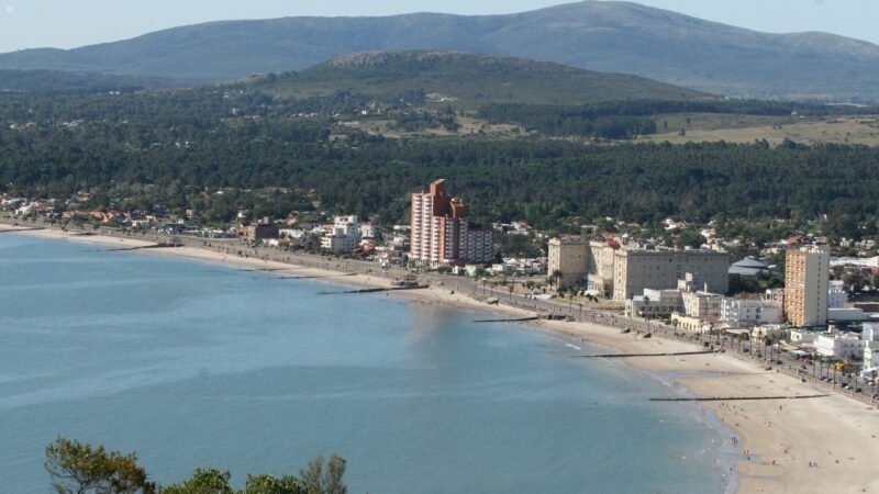 High-angle panorama of a city coastline and sandy beach with a large mountain under a clear blue sky.
