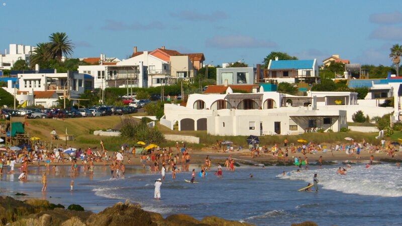 Crowded sandy beach with people in the surf and sun umbrellas in front of white coastal buildings.