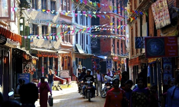 A crowded street with motorbikes and pedestrians under colorful flags, featuring a sign for Tibetan Thanka Production.