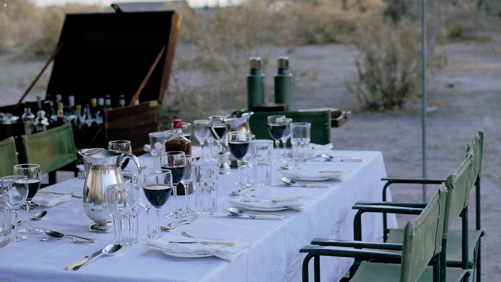 An outdoor dining table laid with wine glasses, cutlery and plates, with white linen tablecloth and khaki cloth-backed chairs