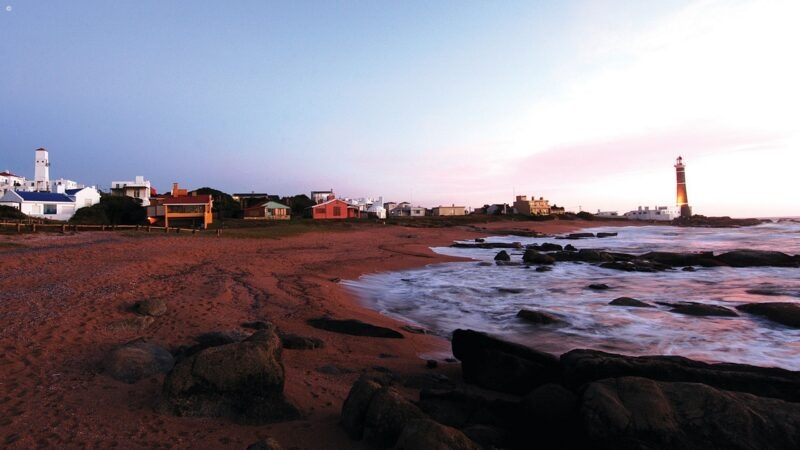 Sunset over a rocky beach with colorful coastal cottages and a distant lighthouse silhouetted against the sky.