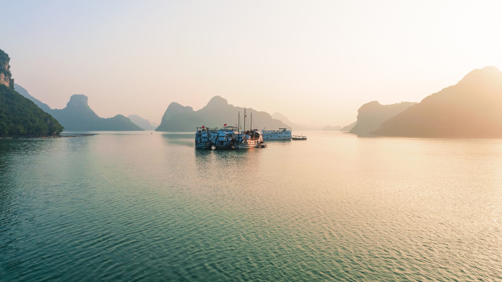Multiple tour boats anchored on a still body of water with large mountain islands in the hazy background.