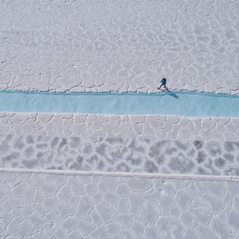 Overhead view of a person in blue walking on a strip of blue water across a vast white textured salt flat. Luxury Argentina tours.