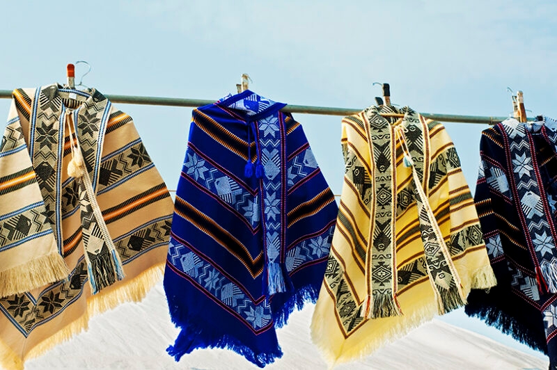 Four brightly colored, patterned ponchos with fringe hanging side-by-side on a metal rod outside. Luxury Argentina tours.