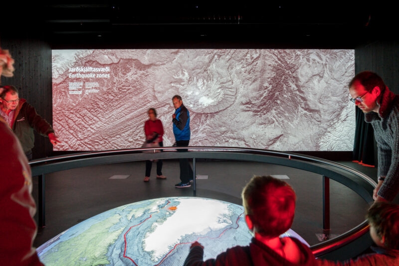 People in a dark museum room view a wall display showing earthquake zones and a projected map of Greenland.