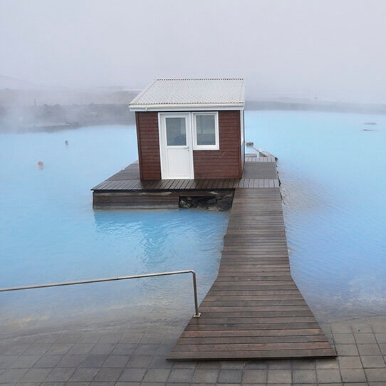 A small brown wooden cabin on a deck is surrounded by the milky blue water of the Blue Lagoon, promoting luxury Iceland tours.