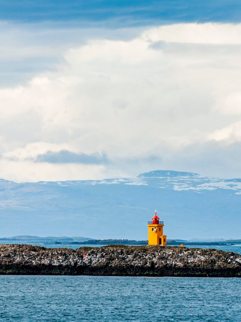 A small, bright yellow lighthouse stands on a dark rocky breakwater