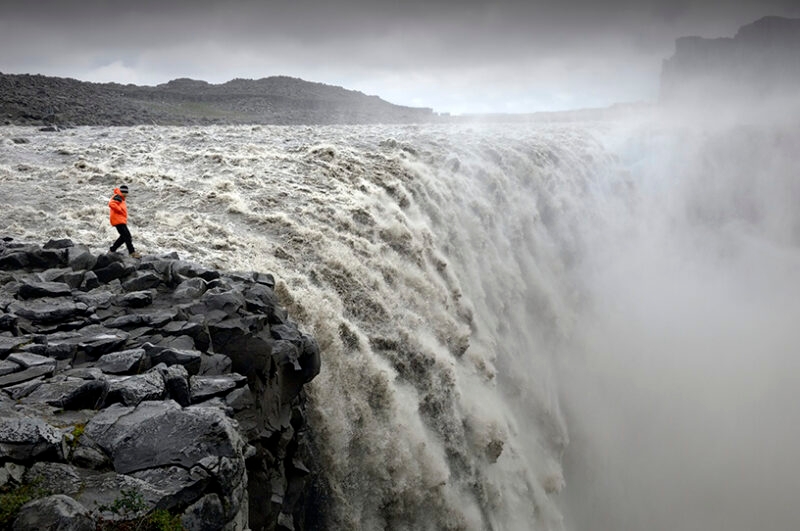 A person in an orange jacket stands on dark rocks at the edge of a massive, misty waterfall, perfect for luxury Iceland trips.