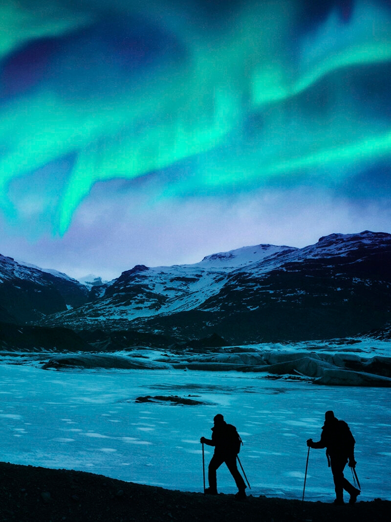 Two hikers stand silhouetted on a frozen glacier under the blue and green Northern Lights, highlighting luxury Iceland tours.