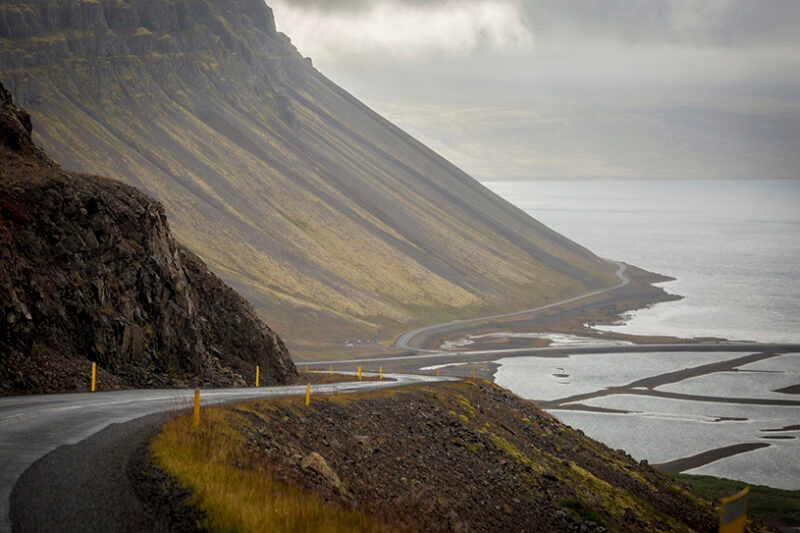 A winding road curves along a steep, grassy coastal mountainside next to water, perfect for luxury Iceland trips.