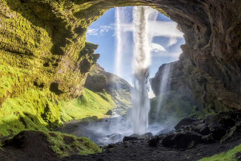 A waterfall streams down from an opening in a mossy green cave into a pool of water, inspiring luxury Iceland vacations.