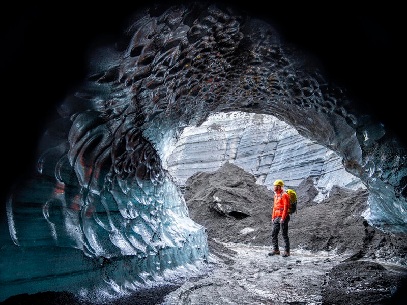 A person in an orange jacket stands inside a sculpted blue ice cave, looking out to the dark glacier, highlighting luxury Iceland tours.