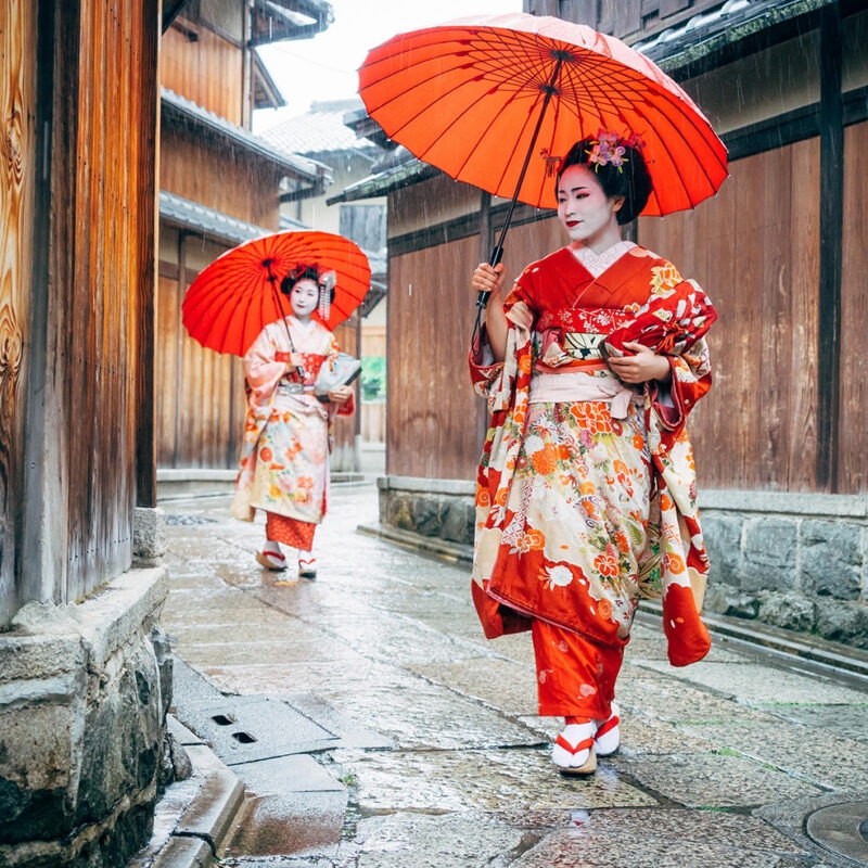 Private Japan Luxury Tours - Maiko Women in traditional kimono clothing holding red parasols Walking in a rainy Kyoto