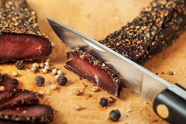 Luxury South African Tours - Close-up of a knife slicing dried beef biltong with peppercorns on a wooden board.