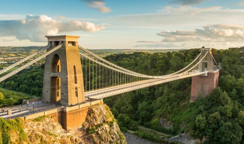The Clifton Suspension Bridge in Bristol, seen from above, spanning a deep, lush, wooded gorge at sunset.