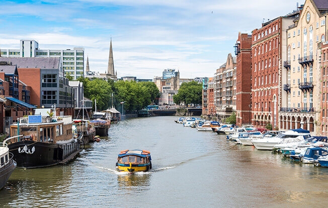View of Bristol's floating harbour with a small yellow and blue boat surrounded by moored boats and city buildings.