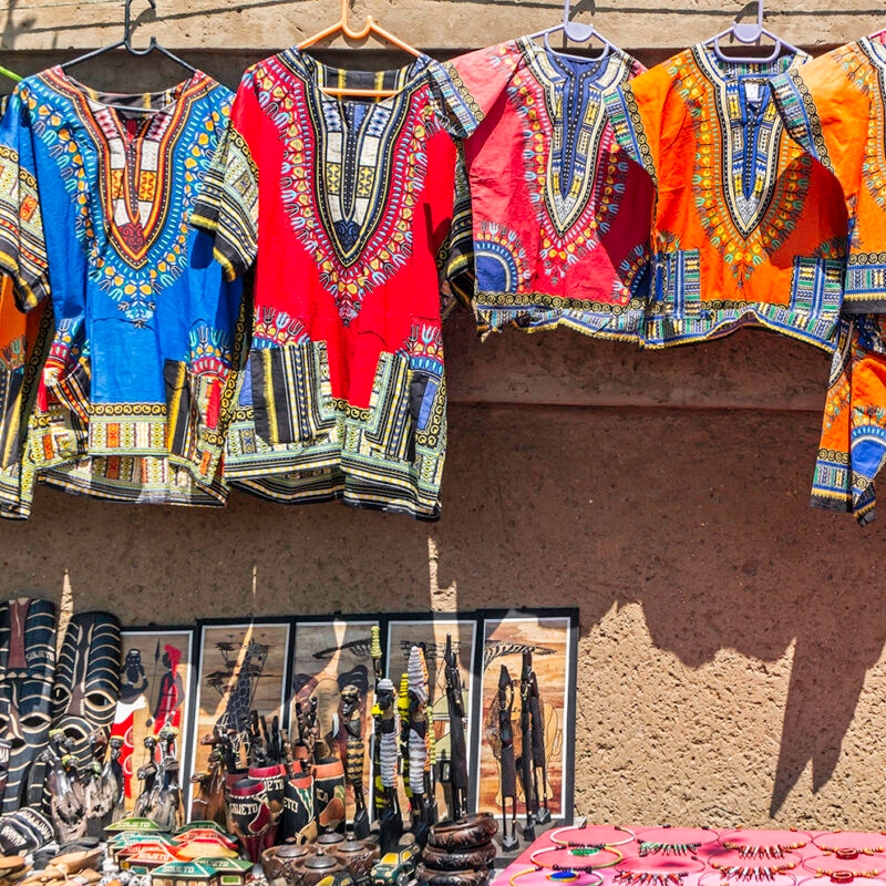 Brightly colored dashiki shirts and various handcrafted African souvenirs displayed for sale at an outdoor market stall.