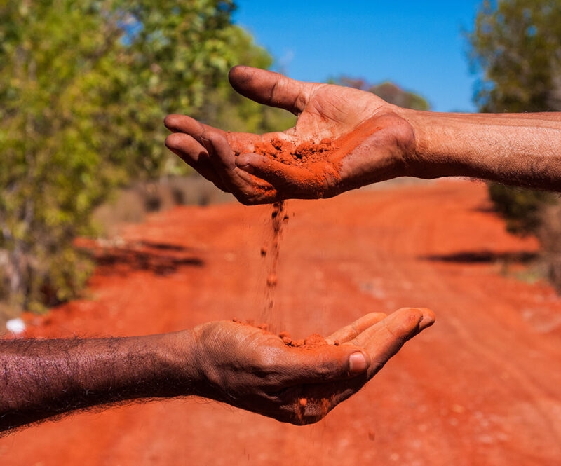 Two pairs of hands hold and pour red dirt/ochre against a backdrop of a reddish outback road, a unique element of luxury Australia vacations.