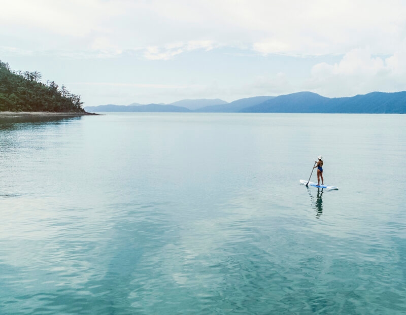 A person stands on a paddleboard in calm, blue-green tropical water with islands in the background, a highlight of luxury Australia trips.
