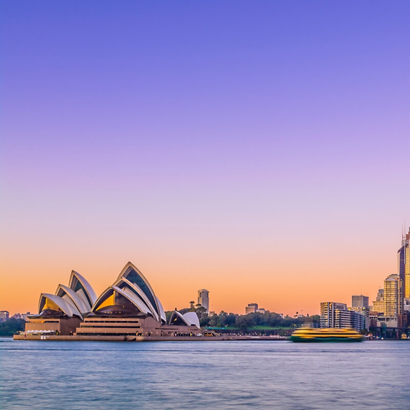 The Sydney Opera House stands by the water at sunset with a cityscape in the background and a purple-orange gradient sky, on luxury Australia tours.