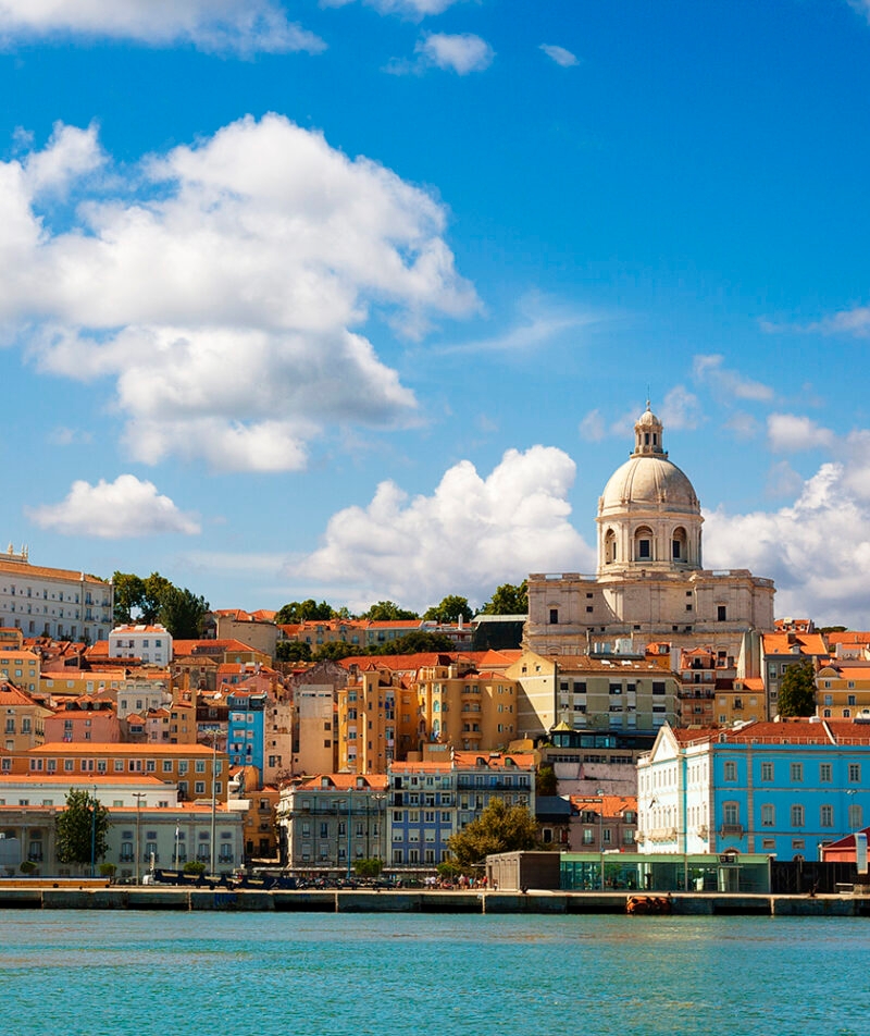 View of Lisbon waterfront with colorful buildings and a large dome church (Santa Engrácia) under a blue sky, luxury Portugal vacations.