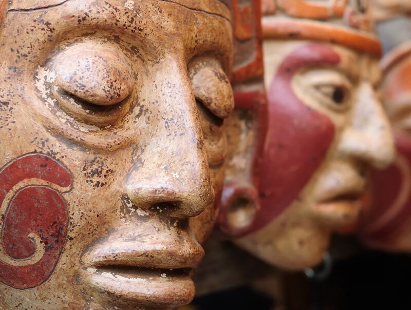 Close-up of multiple hand-crafted clay masks with reddish-brown coloring, featuring closed eyes and red painted accents on the cheeks.