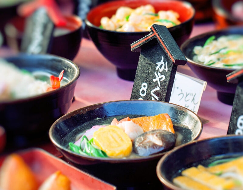 A vibrant display of plastic food models featuring udon bowls with mushrooms and egg, perfect for luxury family tours to Japan.