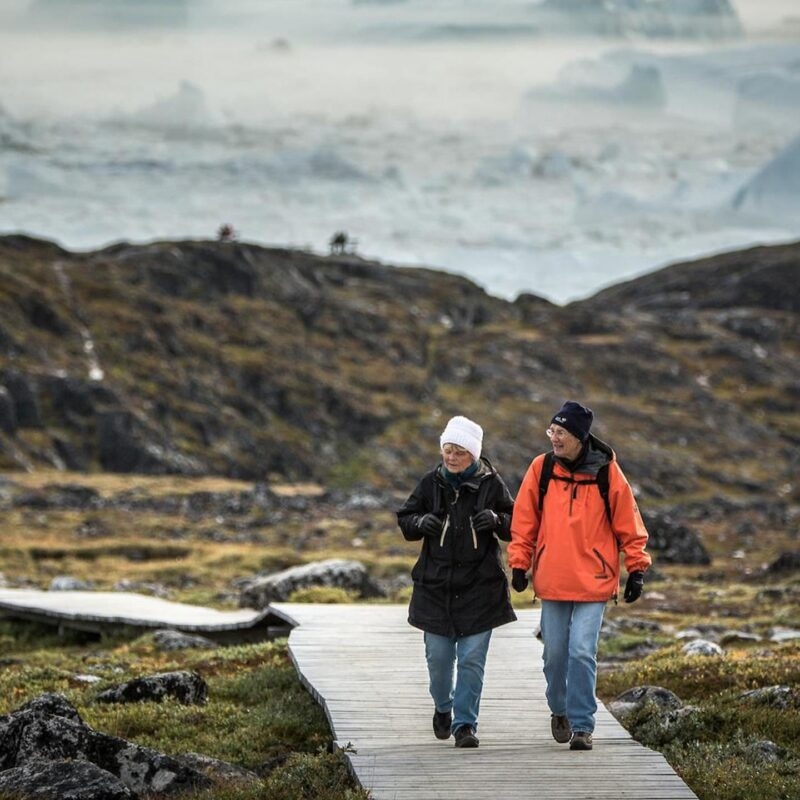 Two people walking on a wooden path through tundra on luxury Polar trips with icebergs behind.