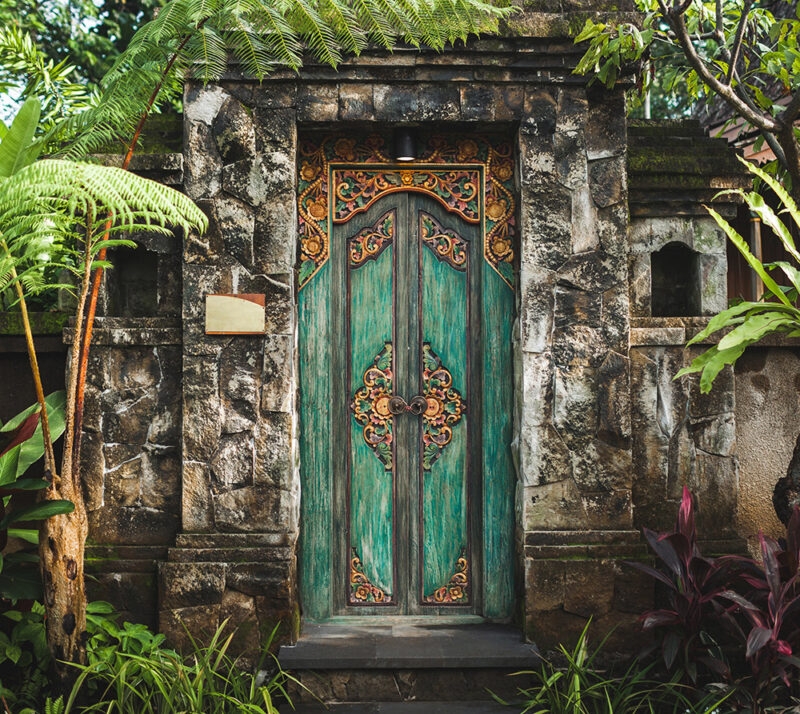 A richly carved, teal and gold double wooden door set in a traditional stone gateway, surrounded by foliage for luxury Indonesia vacations.