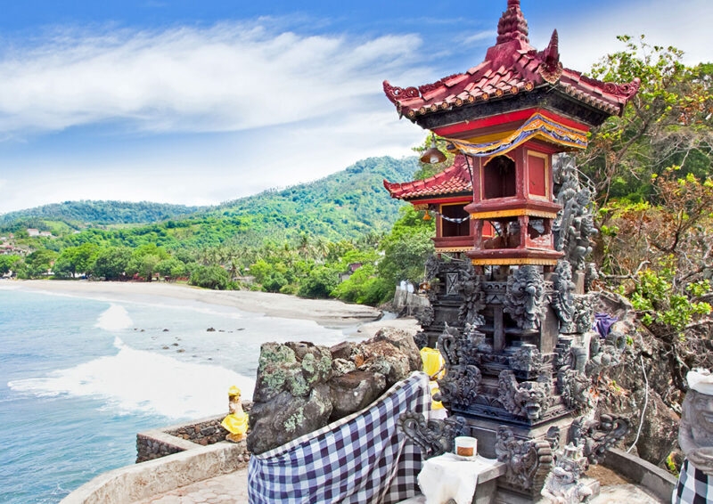 A stone Hindu temple structure on a cliff overlooking a beach and ocean, with lush green hills in the background on luxury Indonesia tours.