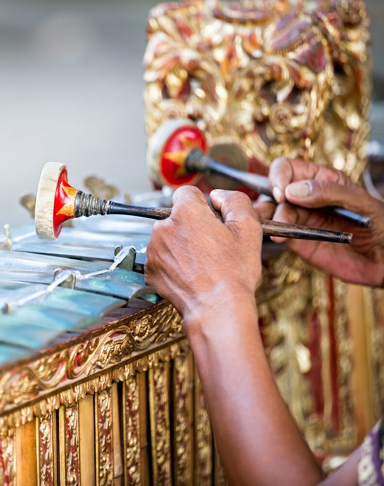 Close-up of hands playing a gamelan instrument with mallets. The ornate, carved wooden frame is gold, highlighting luxury Indonesia trips.
