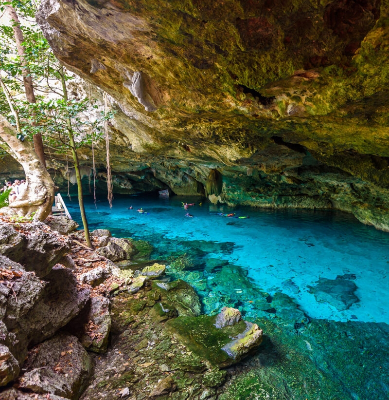 Tourists swim in the clear blue water of a cenote cavern surrounded by rock formations, great for luxury Mexico trips.