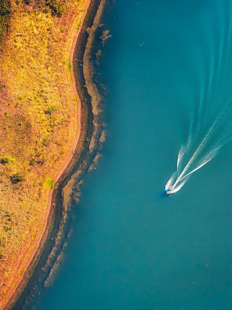 Top-down aerial of a boat wake on blue water by a golden shore during luxury Australasia tours.