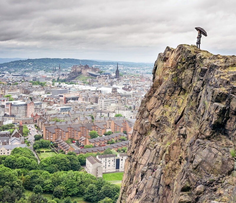 A person stands on the edge of a sheer cliff face overlooking the historic cityscape of Edinburgh and Edinburgh Castle. A highlight of luxury Scotland trips.