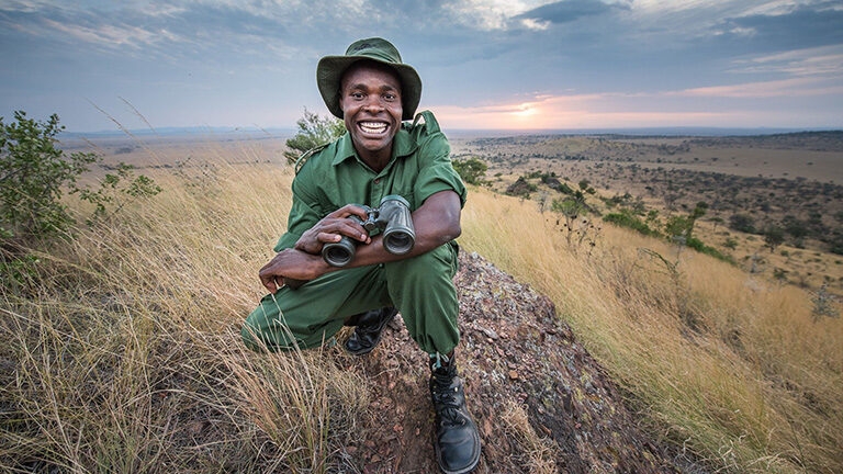 A smiling safari guide holding binoculars on a rocky outcrop during luxury Africa honeymoon tours at sunset.