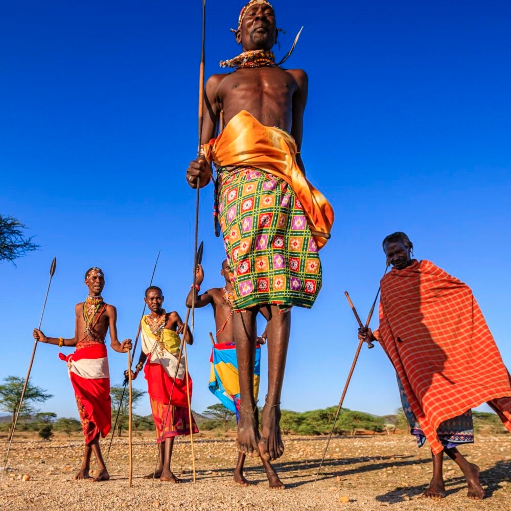 Maasai men in traditional red and patterned clothing performing a jumping dance on luxury Maasai Mara trips.