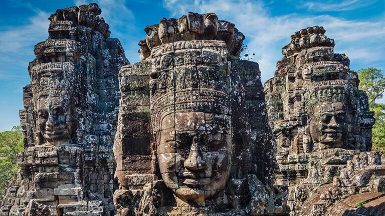 Close-up of three ancient, weathered stone temple towers from Bayon, Angkor Thom, each featuring a colossal, carved, smiling face.