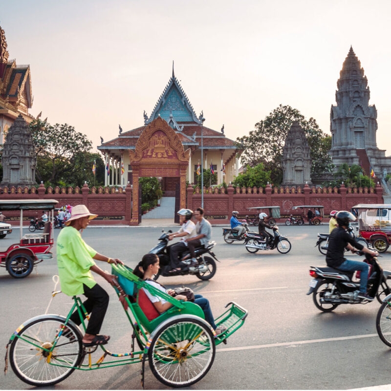 Busy street scene with motorbikes and a cyclo carrying passengers past ornate temple buildings in a Cambodian city.