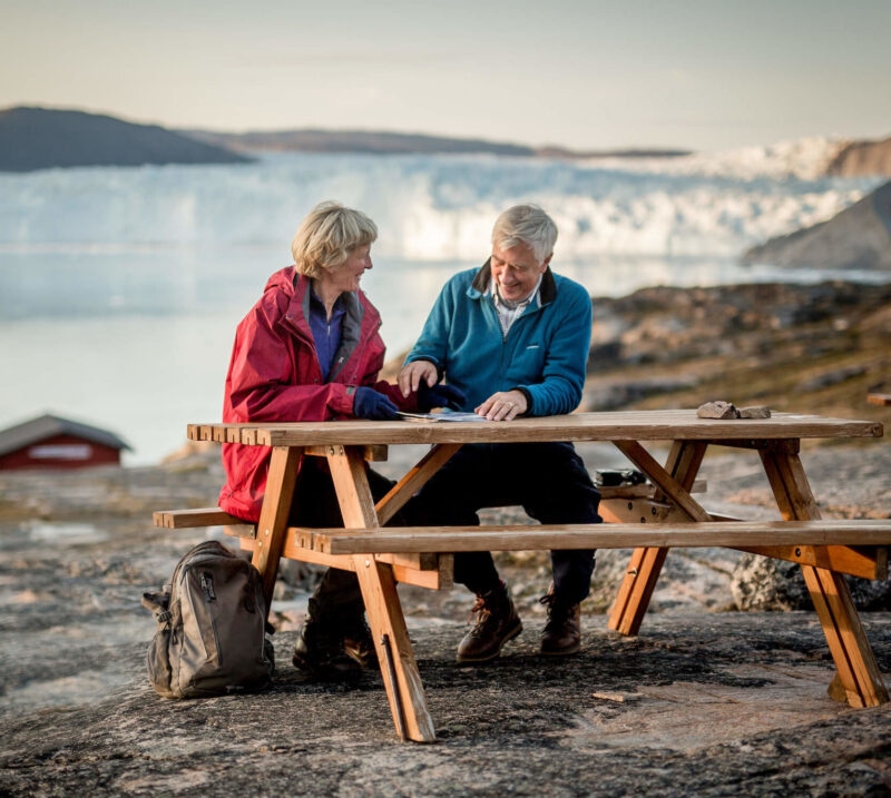 A man and woman sit at a picnic table with a large glacier in the background on luxury Honeymoons.