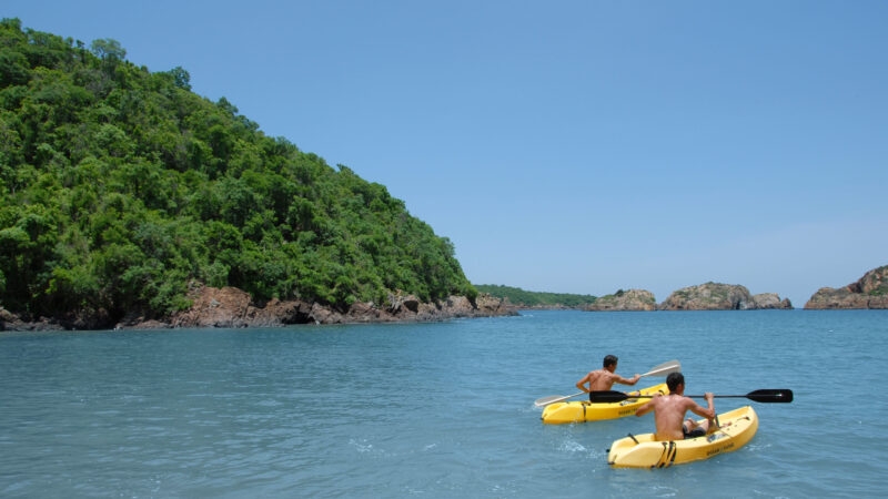 Two people in yellow kayaks paddling in still water by a forested island