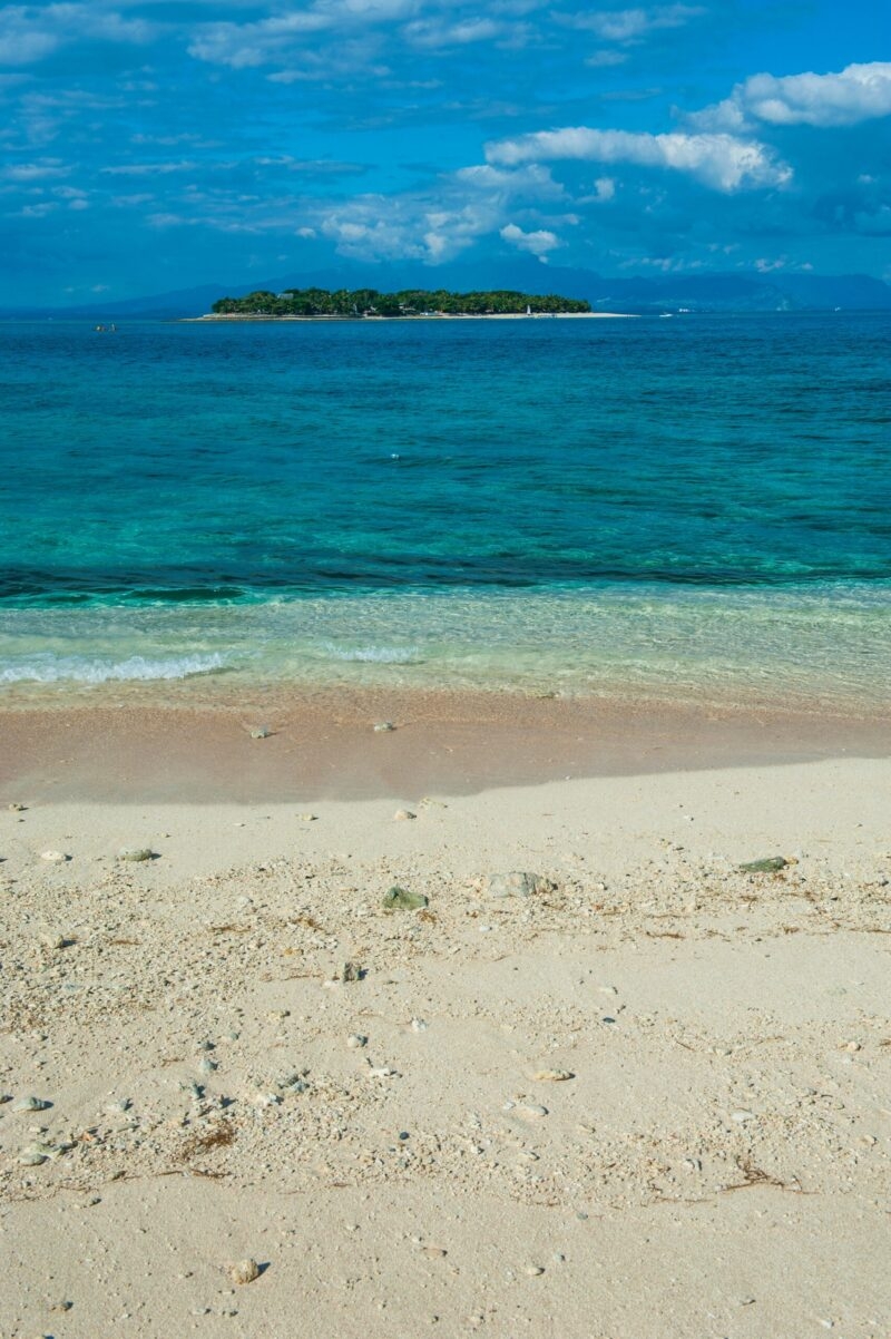 Sandy shoreline looking out over the blue sea toward a small forested island under clouds during luxury Fiji vacations.