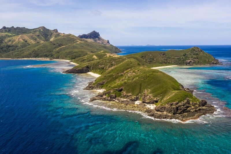 High-angle view of a lush green island surrounded by vibrant coral reefs and blue water for luxury Fiji vacations.