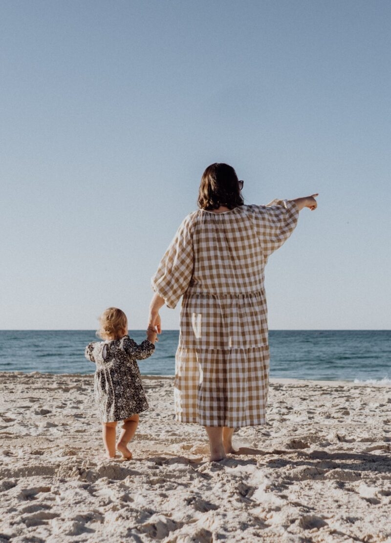 Rear view of a woman and a small child standing on a sandy beach, with the woman pointing toward the blue sea.