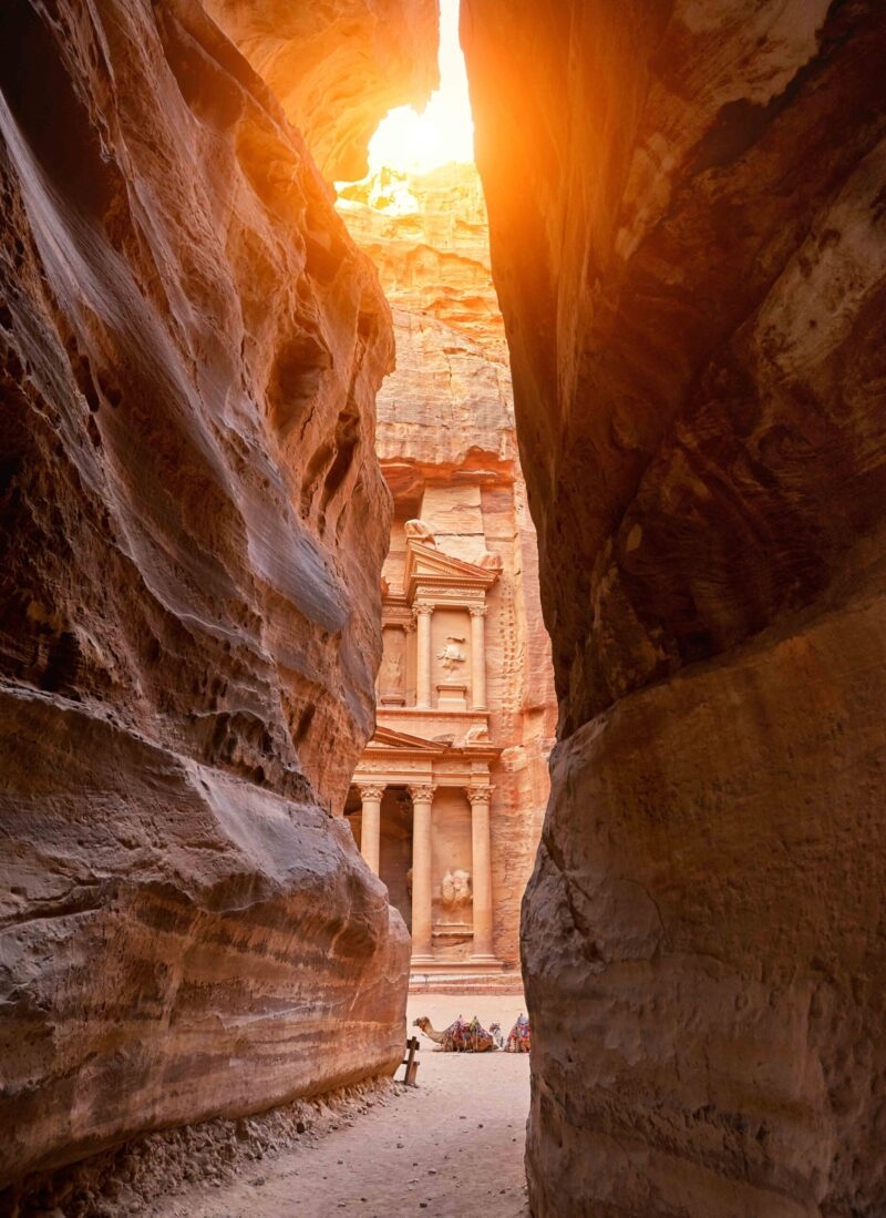 The Treasury (Al-Khazneh) in Petra, Jordan, seen framed through the narrow, high walls of the Siq gorge with bright sun above.