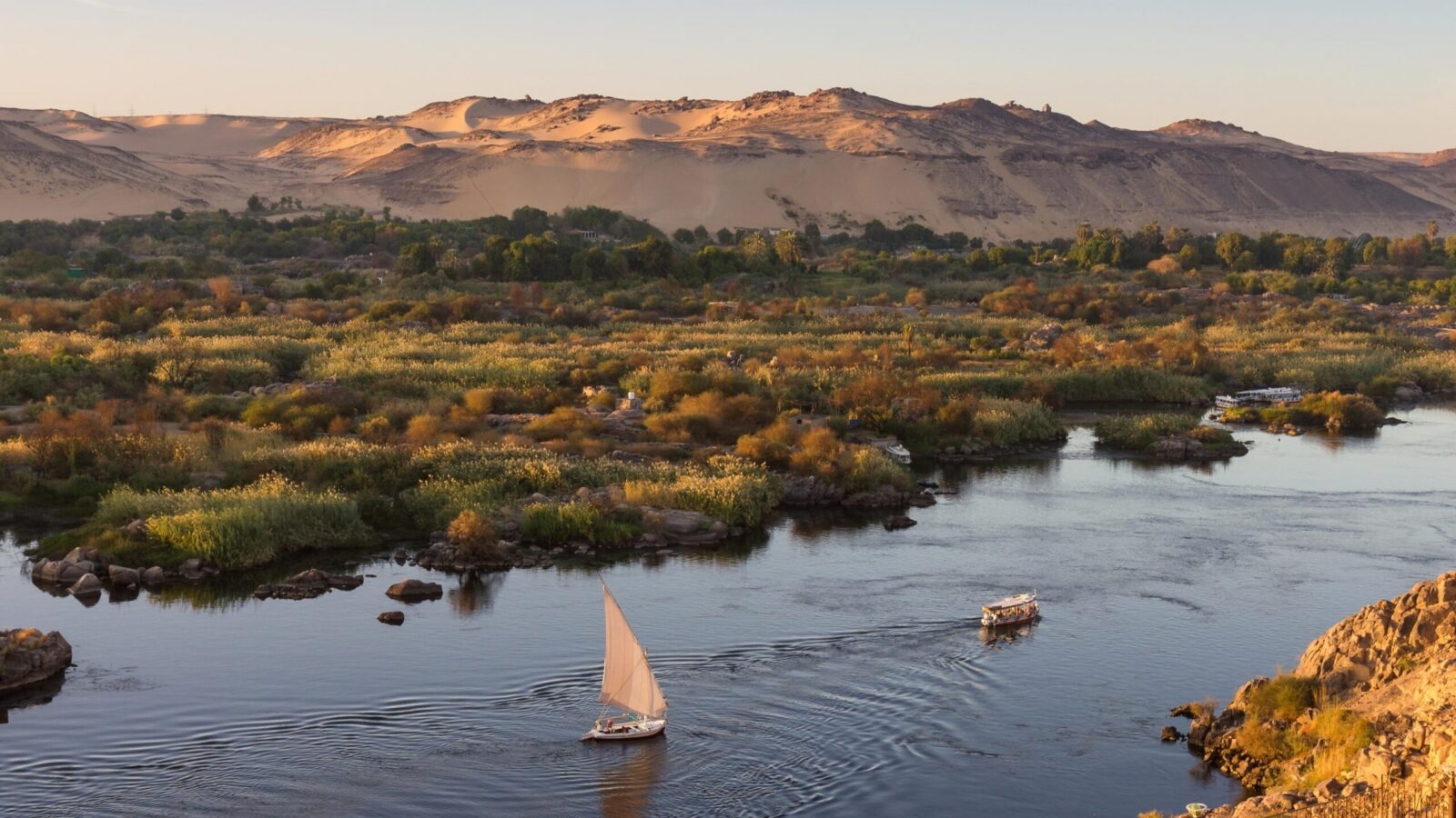 An aerial view of the wide Nile River flowing between green islands and sandy desert hills, with a sailboat, on luxury Middle East trips.